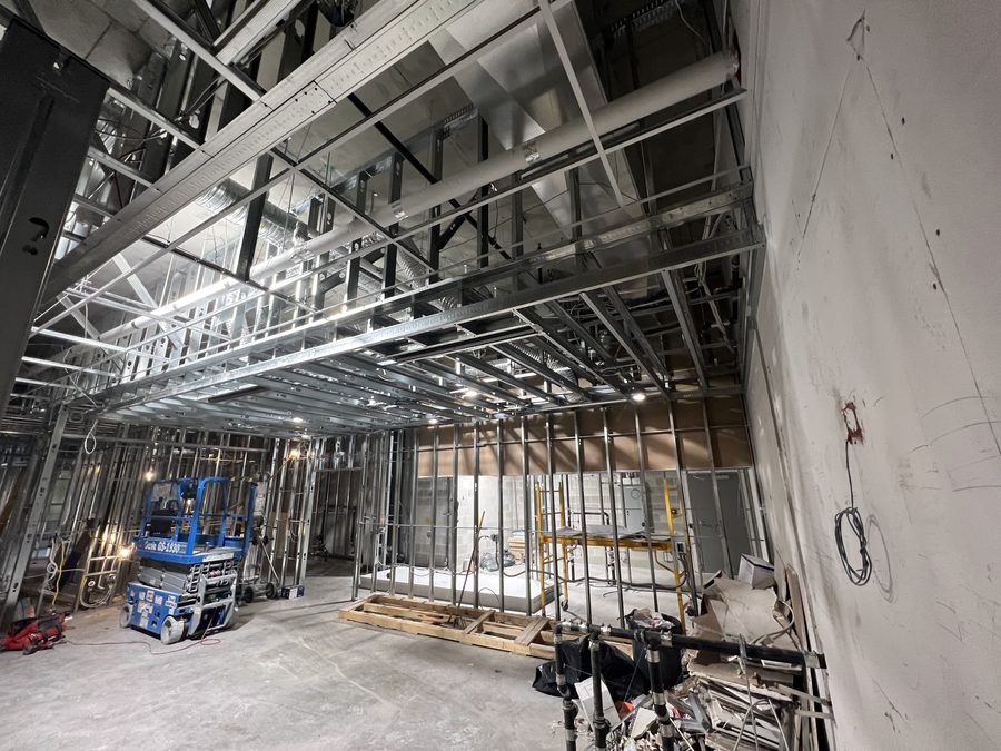 A large open floor with a steel stud ceiling grid and partition framing overhead, a scissor lift below