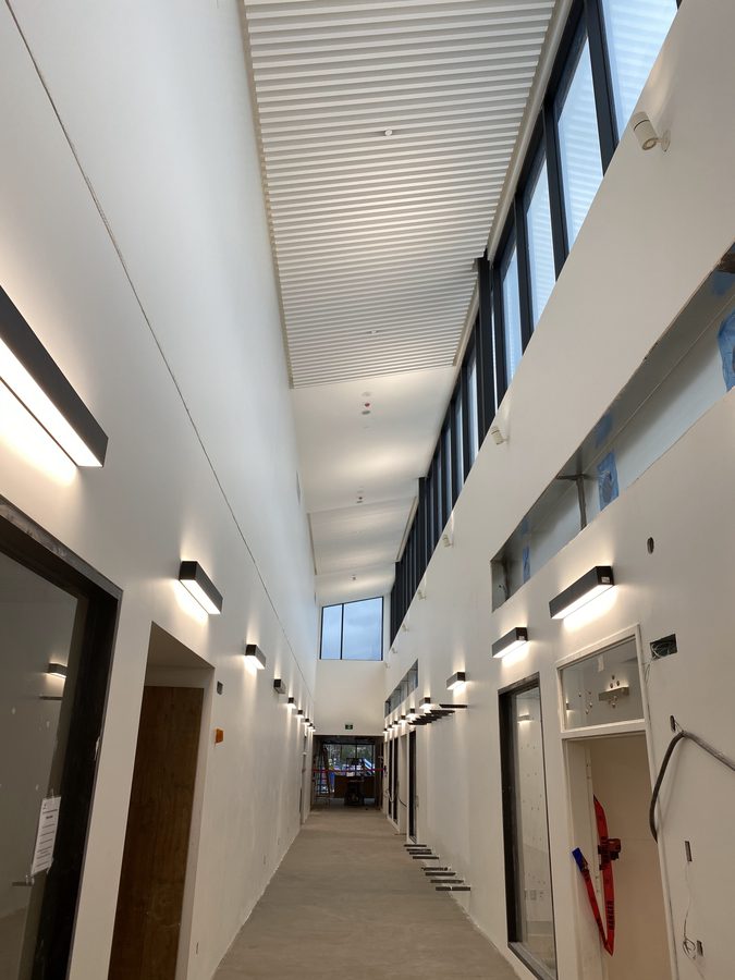 A long finished corridor with clean white walls, wall sconces and a high slatted ceiling under clerestory windows